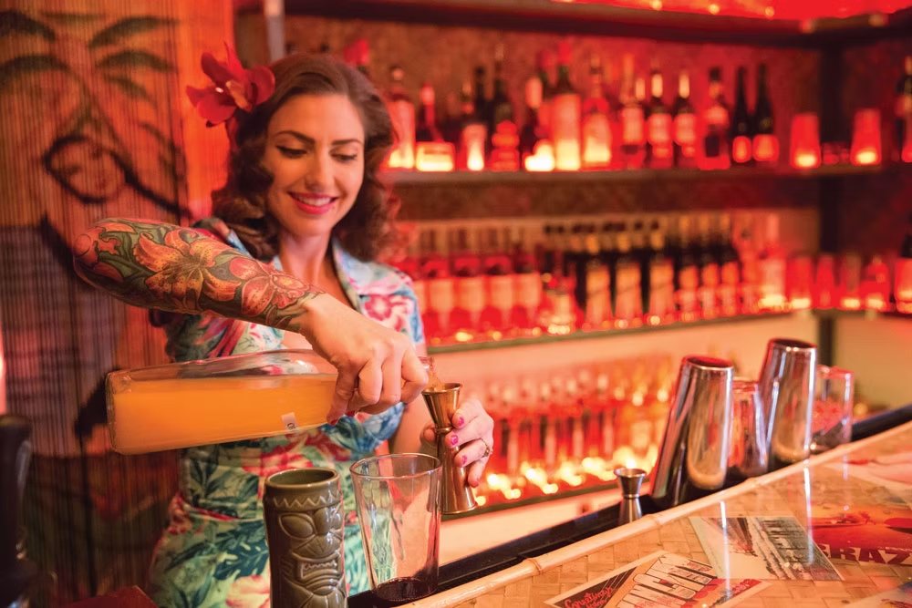 A Bootlegger Tiki bartender in an aloha shirt pouring a cocktail at the back bar